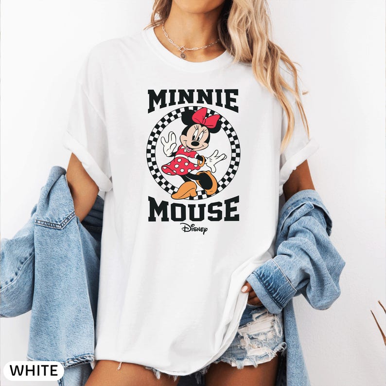 May include: White t-shirt featuring a graphic of Minnie Mouse in a red polka-dot dress, a red bow, and black shoes. The text "MINNIE MOUSE" is above and below the image, with "Disney" below. The shirt is part of a clothing collection.