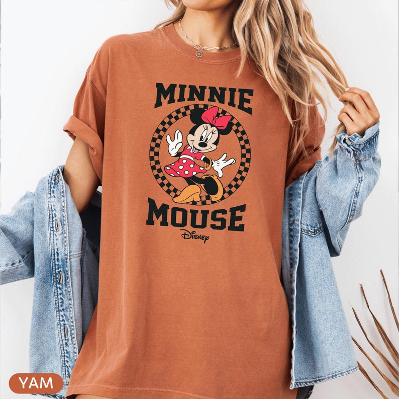 May include: A rust-colored t-shirt featuring a Minnie Mouse graphic. The graphic includes Minnie in a red polka-dot dress and bow, set within a checkered circle, with the words "MINNIE MOUSE" above and below. The Disney logo is also present.