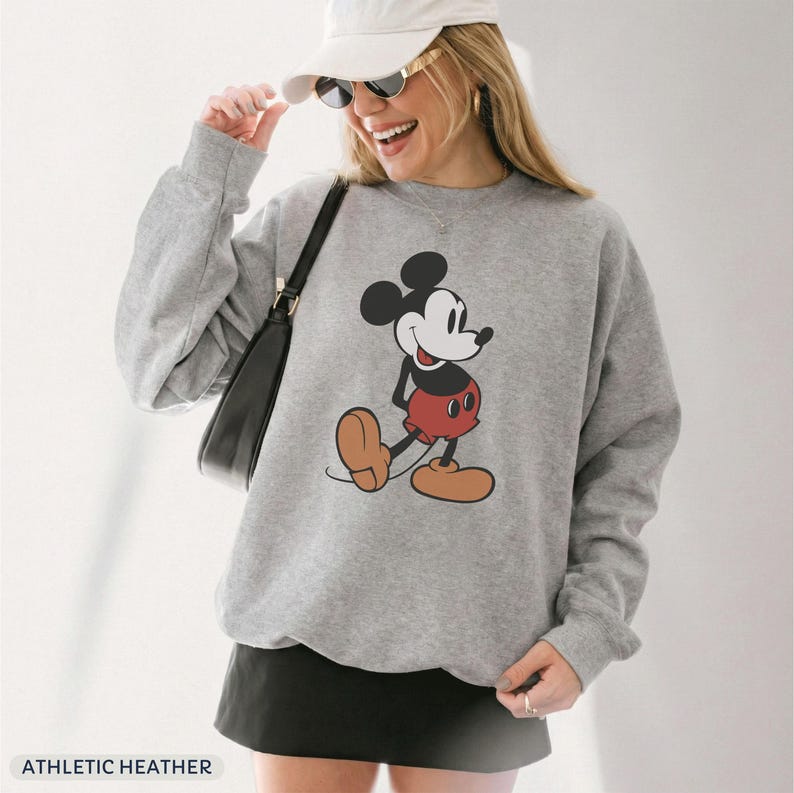 May include: Athletic heather gray sweatshirt featuring a classic Mickey Mouse graphic. The cartoon character is depicted in his iconic pose, wearing red shorts and brown shoes. The sweatshirt has a crew neck and long sleeves. The text "ATHLETIC HEATHER" is at the bottom.