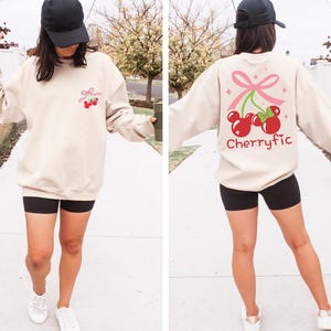 May include: A beige crewneck sweatshirt featuring a small graphic of two red cherries with a pink bow on the left chest. The back displays a larger version of the same cherry graphic with a pink bow and the text Cherryfic in red lettering.