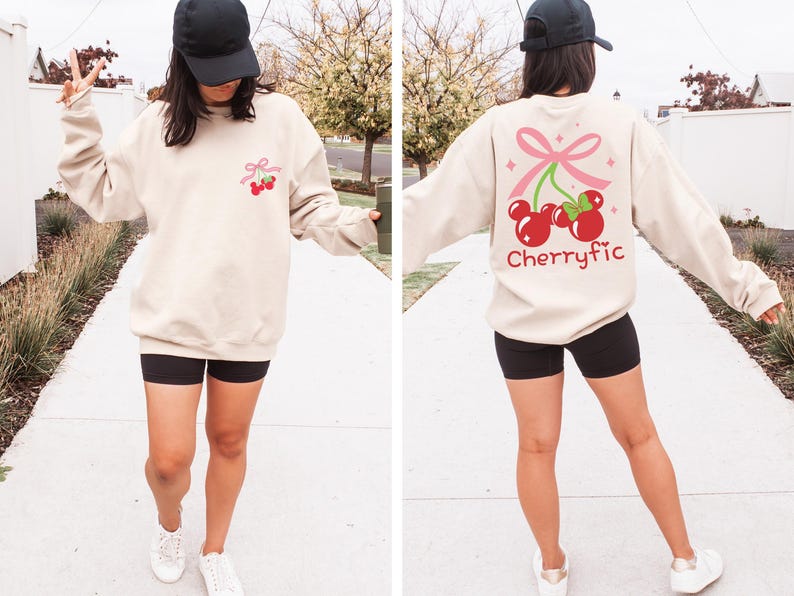 May include: A beige crewneck sweatshirt featuring a small graphic of two red cherries with a pink bow on the left chest. The back displays a larger version of the same cherry graphic with a pink bow and the text Cherryfic in red lettering.