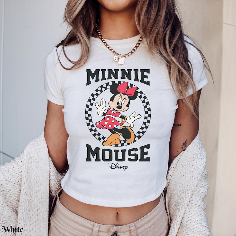 K&ouml;nnte beinhalten: Wei&szlig;es T-Shirt mit einem Minnie Mouse-Motiv. Das Design zeigt Minnie in einem roten gepunkteten Kleid und Schleife, umrahmt von einem schwarz-wei&szlig; karierten Kreis. Die Worte "MINNIE MOUSE" sind oben und unten aufgedruckt.