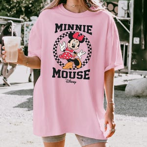 May include: A light pink t-shirt featuring a graphic of Minnie Mouse. The design includes Minnie in a red polka-dot dress, a red bow, and the words "MINNIE MOUSE" in black, with the Disney logo below. The graphic is set within a checkered circle.