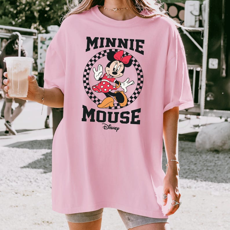 May include: A light pink t-shirt featuring a graphic of Minnie Mouse. The design includes Minnie in a red polka-dot dress, a red bow, and the words "MINNIE MOUSE" in black, with the Disney logo below. The graphic is set within a checkered circle.