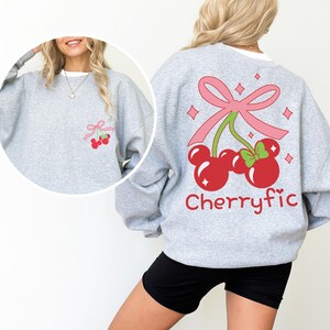 May include: A light gray crewneck sweatshirt featuring a large graphic on the back of two red cherries shaped like mouse ears, tied with a pink bow and accented with sparkles. The word Cherryfic is printed below in red. A smaller version is on the front.