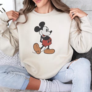 May include: Cream-colored sweatshirt featuring a classic Mickey Mouse graphic. The cartoon character is depicted in his iconic pose, wearing red shorts, brown shoes, and a black and white color scheme. The sweatshirt has long sleeves and a crew neck.