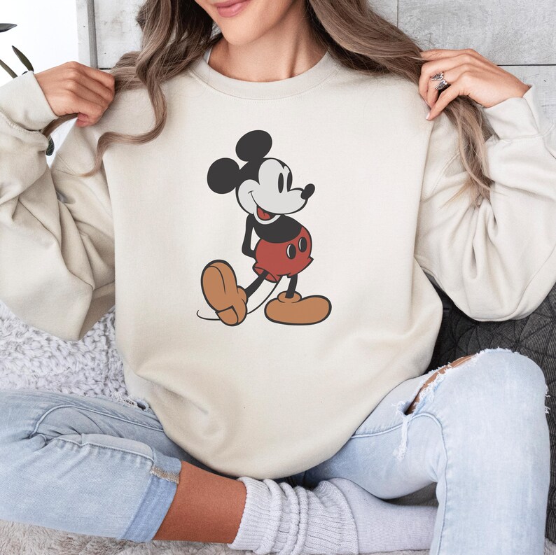 May include: Cream-colored sweatshirt featuring a classic Mickey Mouse graphic. The cartoon character is depicted in his iconic pose, wearing red shorts, brown shoes, and a black and white color scheme. The sweatshirt has long sleeves and a crew neck.