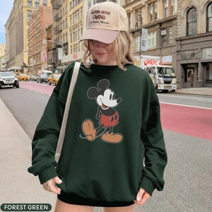 May include: Forest green sweatshirt featuring a classic Mickey Mouse graphic. The cartoon character is depicted in his iconic red shorts, yellow shoes, and white gloves. The sweatshirt is a long-sleeved, casual style.