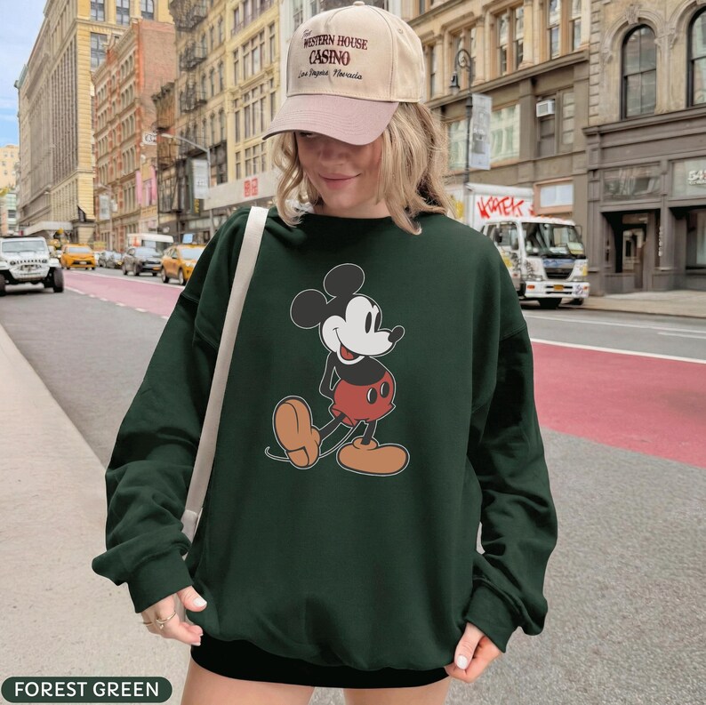 May include: Forest green sweatshirt featuring a classic Mickey Mouse graphic. The cartoon character is depicted in his iconic red shorts, yellow shoes, and white gloves. The sweatshirt is a long-sleeved, casual style.