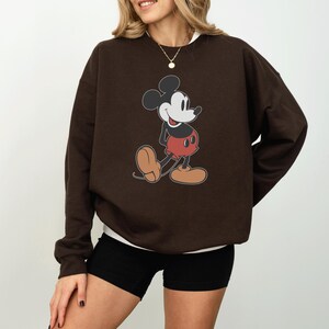 May include: A brown sweatshirt featuring a classic Mickey Mouse graphic. The cartoon character is depicted in his iconic pose, wearing red shorts, black shoes, and white gloves. The sweatshirt is a solid brown color.