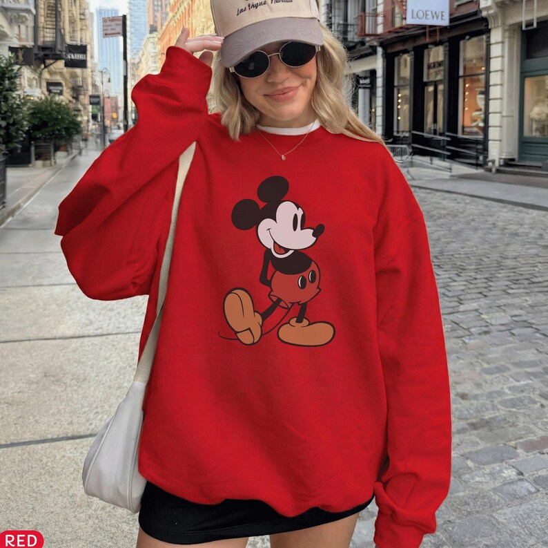 May include: A red sweatshirt featuring a graphic of Mickey Mouse in a walking pose. The sweatshirt has long sleeves and a crew neck. The graphic is in black, white, red, and tan. The word "RED" is in the bottom left corner.