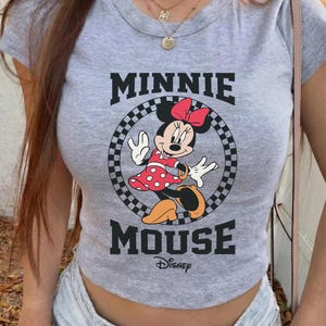 K&ouml;nnte beinhalten: Graues Crop-Top mit Minnie Mouse-Grafik. Das Design zeigt die Worte "MINNIE MOUSE" in schwarzen Buchstaben, mit Minnie Mouse in einem roten Polka-Dot-Kleid und einer roten Schleife. Das Disney-Logo befindet sich darunter.