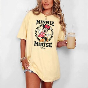 May include: A light yellow t-shirt featuring a graphic of Minnie Mouse. The design includes the text "MINNIE MOUSE" in black, with Minnie Mouse in a red polka dot dress. The shirt is a relaxed fit.