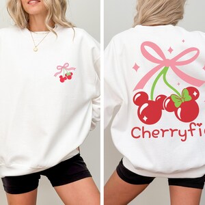 May include: A white long-sleeve sweatshirt featuring a small graphic of two red cherries shaped like mouse heads with a pink bow on the chest. The back displays a larger version of the same cherry graphic with the text Cherryfic in red font.
