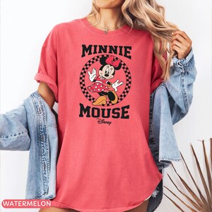May include: A watermelon-colored t-shirt featuring a graphic of Minnie Mouse. The design includes the text "MINNIE MOUSE" in black, with Minnie Mouse in a red polka-dot dress. The Disney logo is below the text.