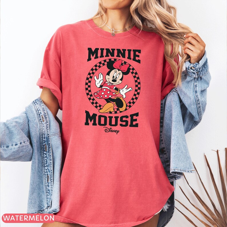 May include: A watermelon-colored t-shirt featuring a graphic of Minnie Mouse. The design includes the text "MINNIE MOUSE" in black, with Minnie Mouse in a red polka-dot dress. The Disney logo is below the text.