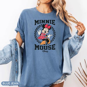May include: Blue jean t-shirt featuring a graphic of Minnie Mouse in a red polka dot dress, a red bow, and black shoes. The text "MINNIE MOUSE" is above and "Disney" is below the graphic. The shirt has rolled-up sleeves.
