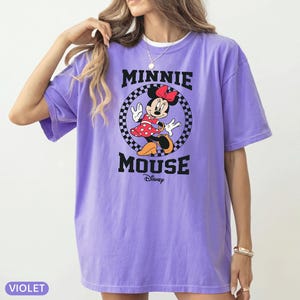 May include: Violet-colored t-shirt featuring a graphic of Minnie Mouse. The design includes Minnie in a red polka-dot dress, a black and white checkered circle, and the words "MINNIE MOUSE" in black block letters. The Disney logo is below.