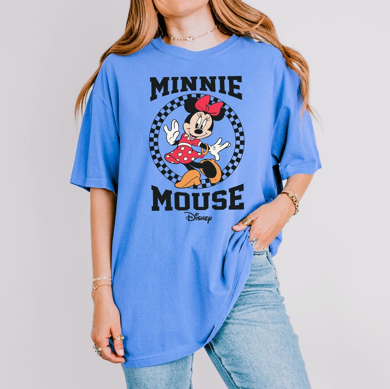 May include: A light blue t-shirt featuring a graphic of Minnie Mouse in a red polka dot dress, a red bow, and black shoes. The words "MINNIE MOUSE" are printed above and below the character in black, with the Disney logo below.