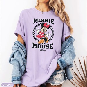 May include: Orchid-colored t-shirt featuring a Minnie Mouse graphic. The graphic includes Minnie Mouse in a red polka-dot dress, a black and white checkered circle, and the words "MINNIE MOUSE" and "Disney" in black.