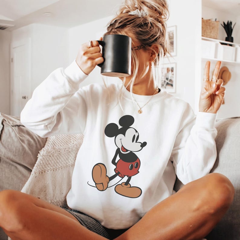 May include: White sweatshirt featuring a classic Mickey Mouse graphic. The cartoon character is depicted in black, red, and tan colors. The sweatshirt has long sleeves and a crew neck. A black mug is held up to the face.