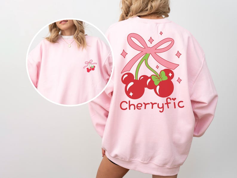 May include: A light pink crewneck sweatshirt featuring a large graphic on the back of two red cherries shaped like mouse ears, tied with a pink ribbon bow, and the text Cherryfic. A smaller version of the same cherry graphic is on the front left chest.
