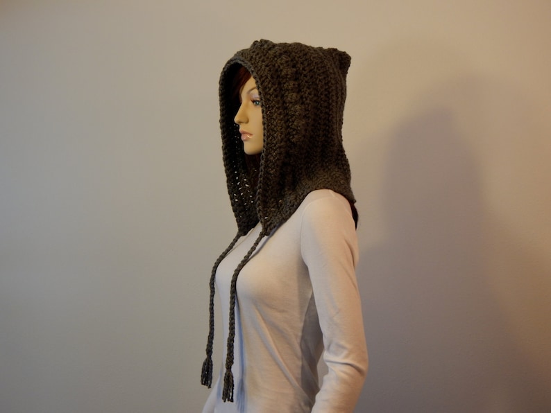May include: A dark olive green crocheted hood with ear-like points at the top. The hood has long, braided ties with tassels. The hood is displayed on a mannequin, showcasing the handmade texture and design.