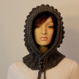May include: A gray crocheted hood with a textured, scalloped edge. The hood features a neck warmer with two large, dark buttons and a drawstring. The handmade hood is displayed on a mannequin.