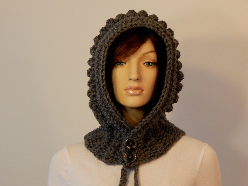 May include: A gray crocheted hood with a textured, scalloped edge. The hood features a neck warmer with two large, dark buttons and a drawstring. The handmade hood is displayed on a mannequin.