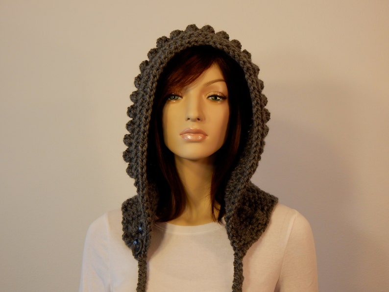 May include: A gray, handmade crochet hood with a textured, scalloped edge. The hood features two buttons and long ties. The hood is designed to be worn over the head and shoulders.
