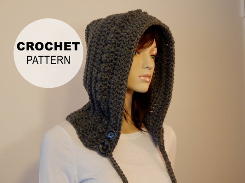 May include: A dark gray crocheted hood with ear flaps and two large buttons. The hood is displayed on a mannequin. The text "CROCHET PATTERN" is in a white circle in the upper left corner.