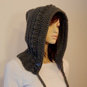 May include: A gray crocheted hood with ear-like points at the top. The hood features a button closure at the neck and long, braided ties. The handmade hood is displayed on a mannequin, showcasing its texture and design. The hood is a cozy accessory.