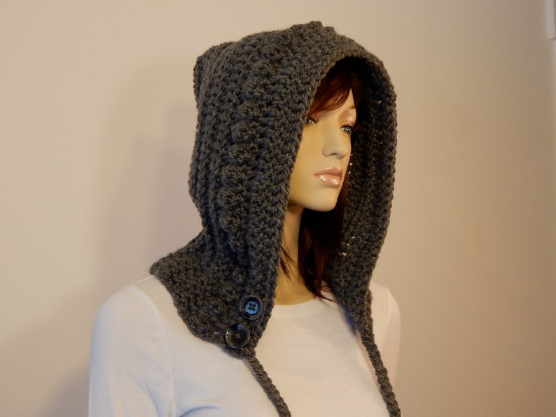 May include: A gray crocheted hood with ear-like points at the top. The hood features a button closure at the neck and long, braided ties. The handmade hood is displayed on a mannequin, showcasing its texture and design. The hood is a cozy accessory.