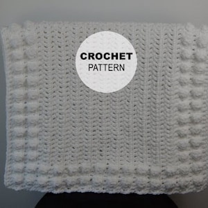 May include: White crochet baby blanket with a textured stitch pattern and a border of raised bobbles. The blanket has a white circle label with the text "CROCHET PATTERN" in black.