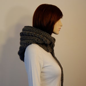 May include: A gray crocheted cowl scarf with a hood. The scarf is made of thick yarn and has a textured appearance. The hood is designed to cover the head and neck, with a long, dangling section.