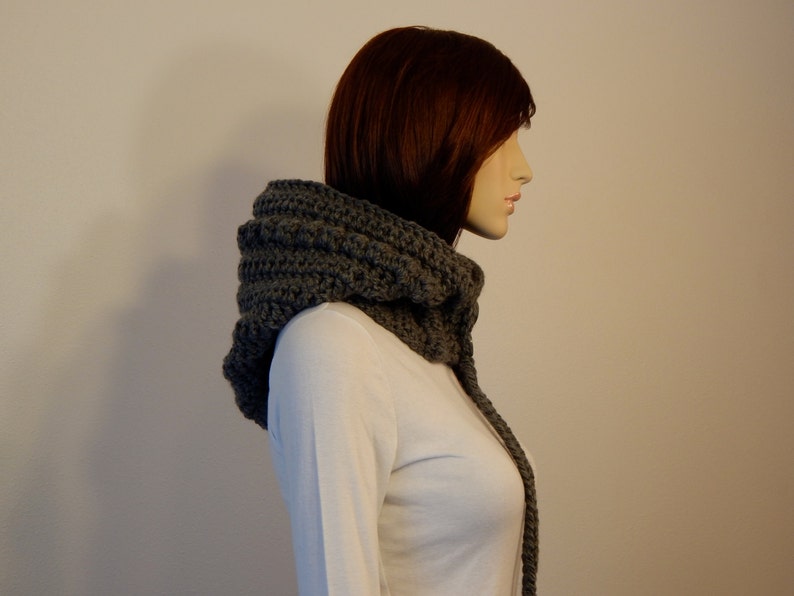 May include: A gray crocheted cowl scarf with a hood. The scarf is made of thick yarn and has a textured appearance. The hood is designed to cover the head and neck, with a long, dangling section.