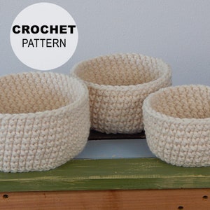 May include: Three crocheted baskets in various sizes, all in a cream color. The baskets are sitting on a wooden surface with a green top.