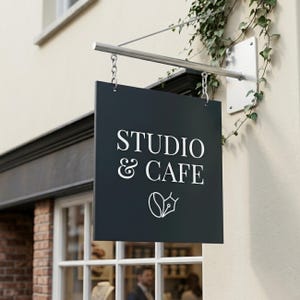 May include: A square black hanging sign with white text that reads STUDIO & CAFE above a graphic icon of a coffee bean combined with a fountain pen nib. The sign is suspended by two silver chains from a horizontal metal bar attached to a wall.