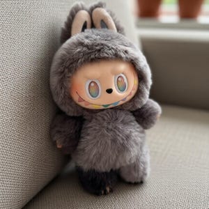 May include: A plush doll in a grey furry bunny suit with a smiling face and rainbow details. The doll has a hood with bunny ears and is sitting on a grey sofa.