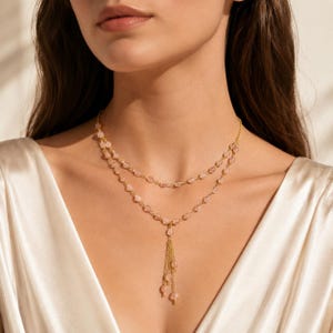 May include: A double-layered gold chain necklace featuring small, translucent, pale pink teardrop-shaped beads spaced along the links. The necklace includes a central drop element consisting of multiple short, dangling gold chains tipped with pink beads.