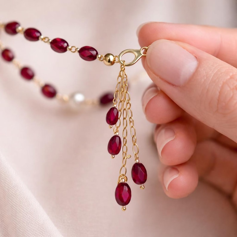 May include: A gold chain necklace featuring small, oval-shaped, deep red faceted beads spaced along the links. A gold lobster clasp connects to a dangling charm cluster composed of four delicate gold chains, each ending in a single red bead.