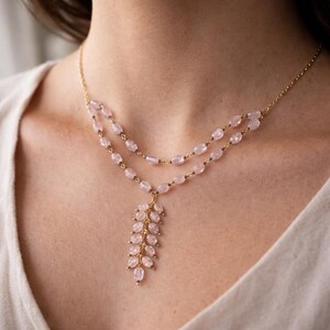 May include: A double-strand necklace featuring small, oval-shaped, pale pink stones linked by a thin gold-tone chain. The necklace includes a central pendant composed of multiple pink stone beads arranged in a vertical, leaf-like cluster pattern.