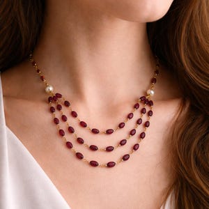 May include: A three-strand necklace featuring small, oval-shaped deep red beads linked by a thin gold-tone chain. The necklace includes two white spherical pearls positioned symmetrically on each side, connecting the strands to the main chain.