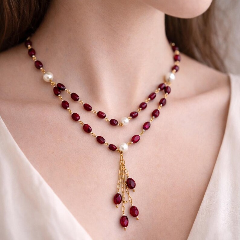 May include: A double-strand necklace featuring deep red oval beads and white round pearls linked by a thin gold-tone chain. The center of the necklace includes a dangling tassel element composed of several gold-tone chains with red beads.