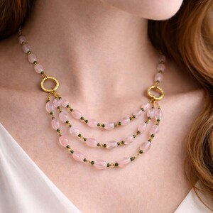 May include: A multi-strand necklace featuring oval-shaped pale pink beads and small dark green accent beads. The necklace is constructed with gold-toned metal links and two textured gold-toned circular connectors that join the three draped strands.