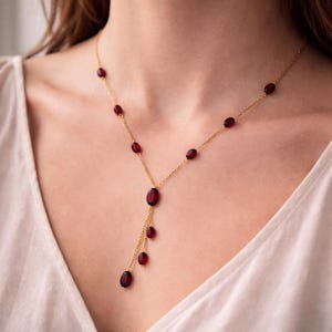 May include: A gold chain necklace featuring multiple small, smooth, oval-shaped dark red stones spaced along the length of the chain. The necklace includes a central drop section with three additional dangling oval red stones at the bottom.