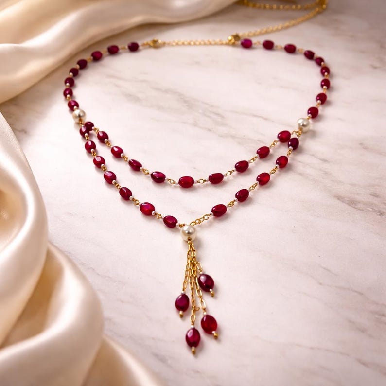 May include: A double-strand necklace featuring small, oval-shaped deep red beads and white spherical beads linked by a thin gold-tone chain. The design includes a central drop pendant composed of multiple gold-tone chains tipped with red beads.