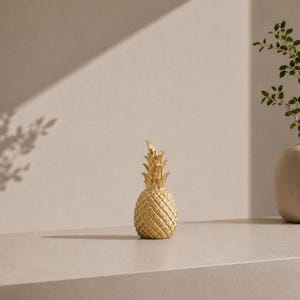 May include: A metallic gold pineapple figurine featuring a textured, diamond-patterned body and a pointed, leafy crown. The object is rendered in a solid, reflective gold finish, capturing the distinct shape and segmented exterior of a pineapple.