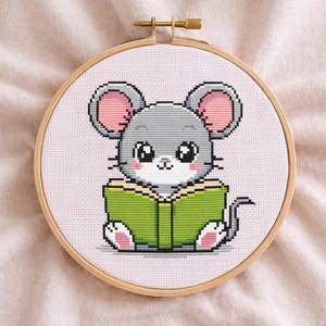 May include: Cross-stitch embroidery of a gray mouse reading a green book, set in a wooden hoop. The mouse has pink ears and paws, and large, expressive eyes. The background is a light pink fabric.