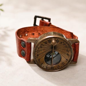 May include: A wrist-worn sundial featuring a circular brass face with engraved numbers and symbols, attached to a textured reddish-brown leather strap. The strap is secured with a dark metal buckle and features two visible metal rivets on the side.